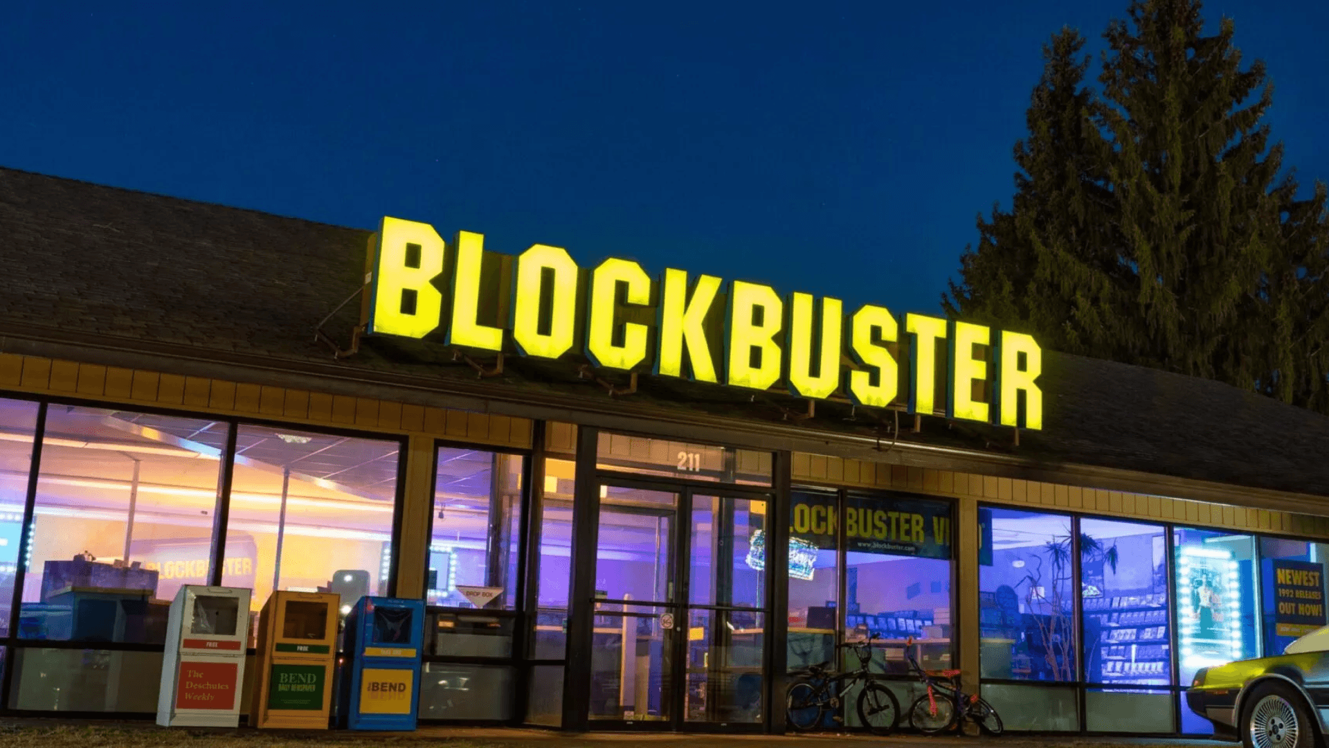 Night photograph of a Blockbuster Video store with iconic yellow illuminated signage glowing against a dark evening sky with pine trees, representing the famous retail video chain that failed to adapt to digital disruption