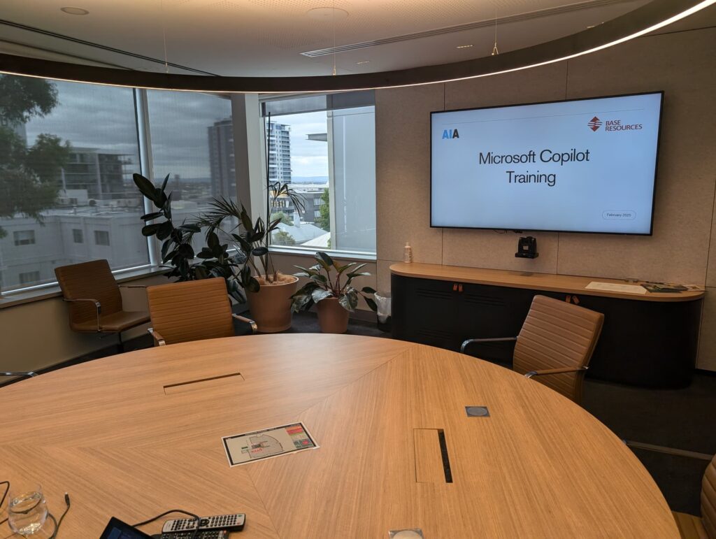 Executive boardroom with round wooden table and city views showing Microsoft Copilot Training presentation screen with AI Advancements and Base Resources logos, representing corporate AI training delivery